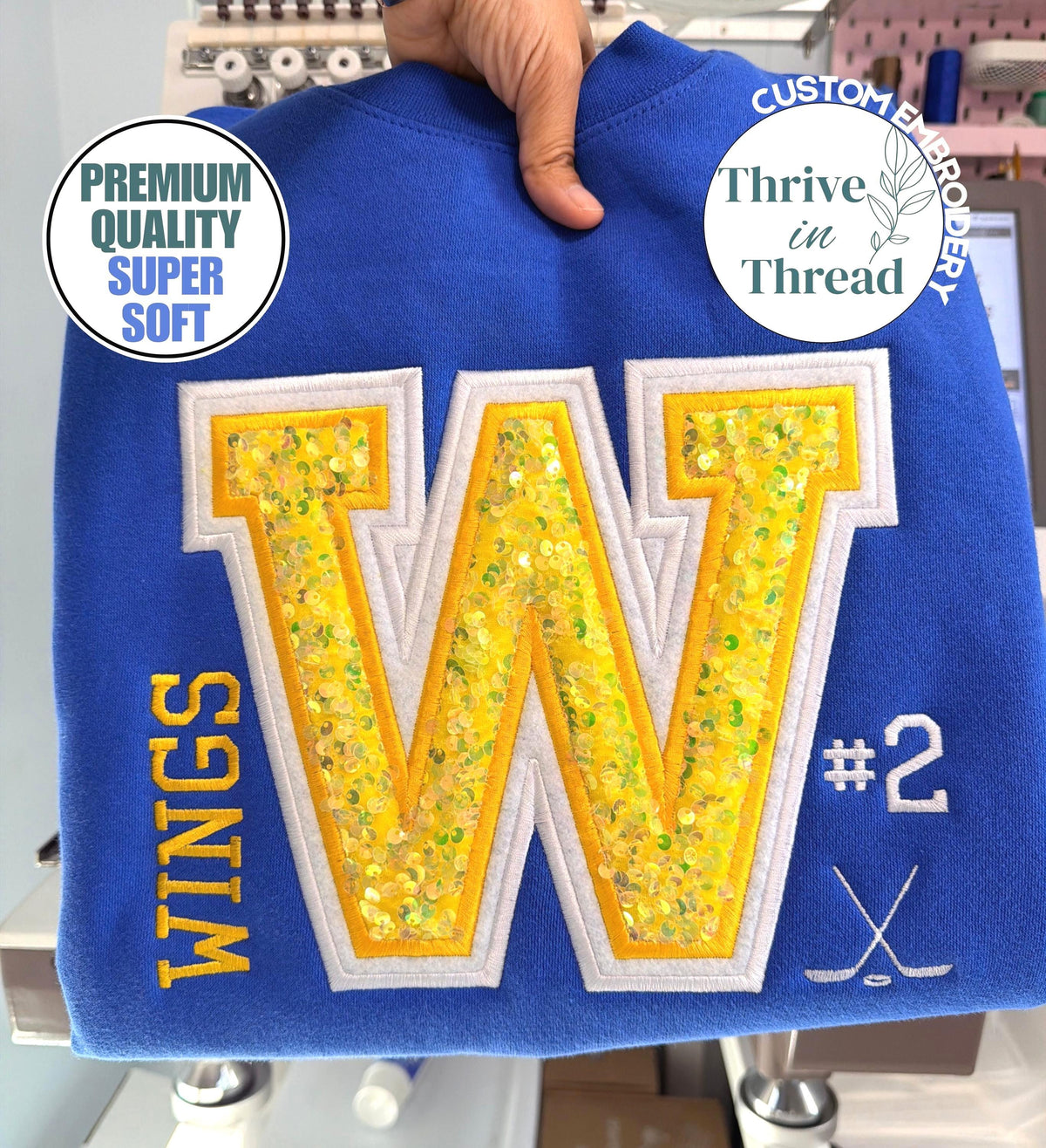 May include: A royal blue sweatshirt featuring a large central letter W with yellow sequin fill and white embroidery outline. The word WINGS is embroidered in yellow on the left, with the number #2 and a crossed hockey stick icon in white on the right.