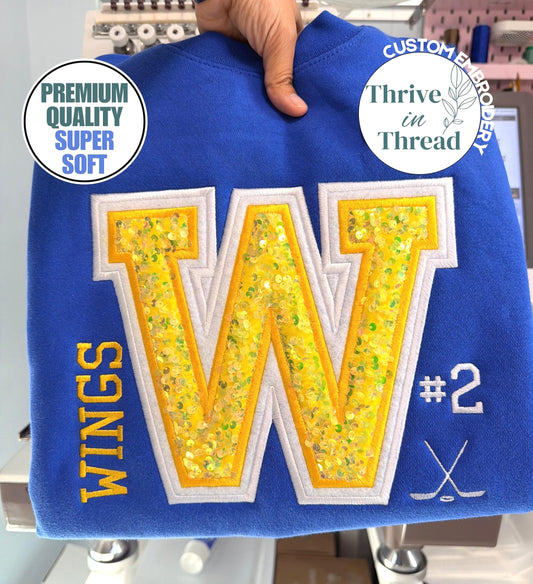 May include: A royal blue sweatshirt featuring a large central letter W with yellow sequin fill and white embroidery outline. The word WINGS is embroidered in yellow on the left, with the number #2 and a crossed hockey stick icon in white on the right.